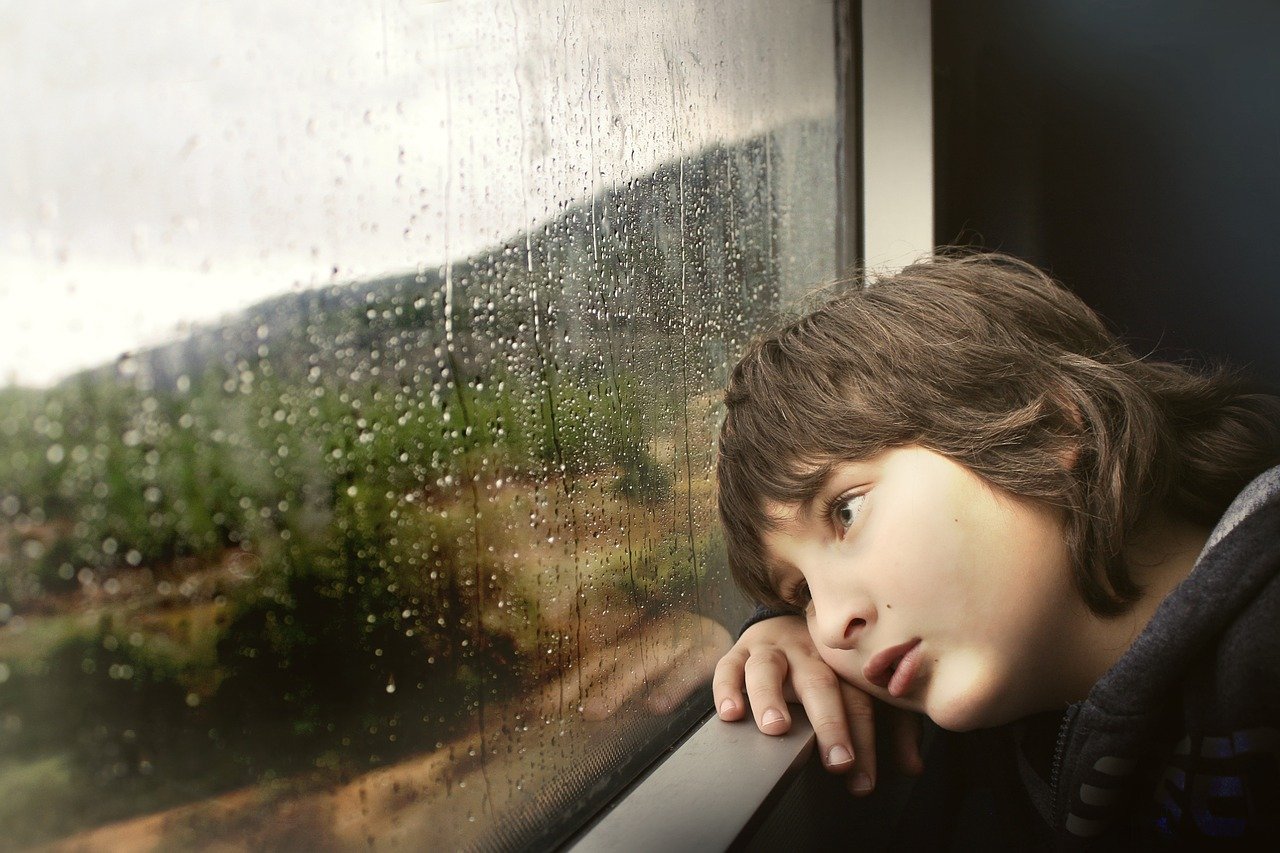 child looks longingly through a rain covered window; remote learning concept