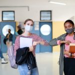 Young students friends with face masks back at college or university greeting, coronavirus concept.