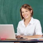 Happy woman typing on a laptop sitting at a table in front of green chalkboard smiling at the camera