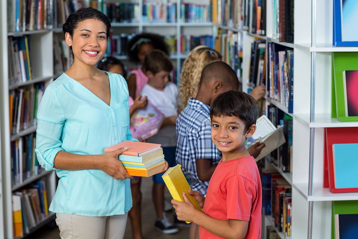 Portrait smiling teacher giving books to a boy in library; book report lesson plans concept