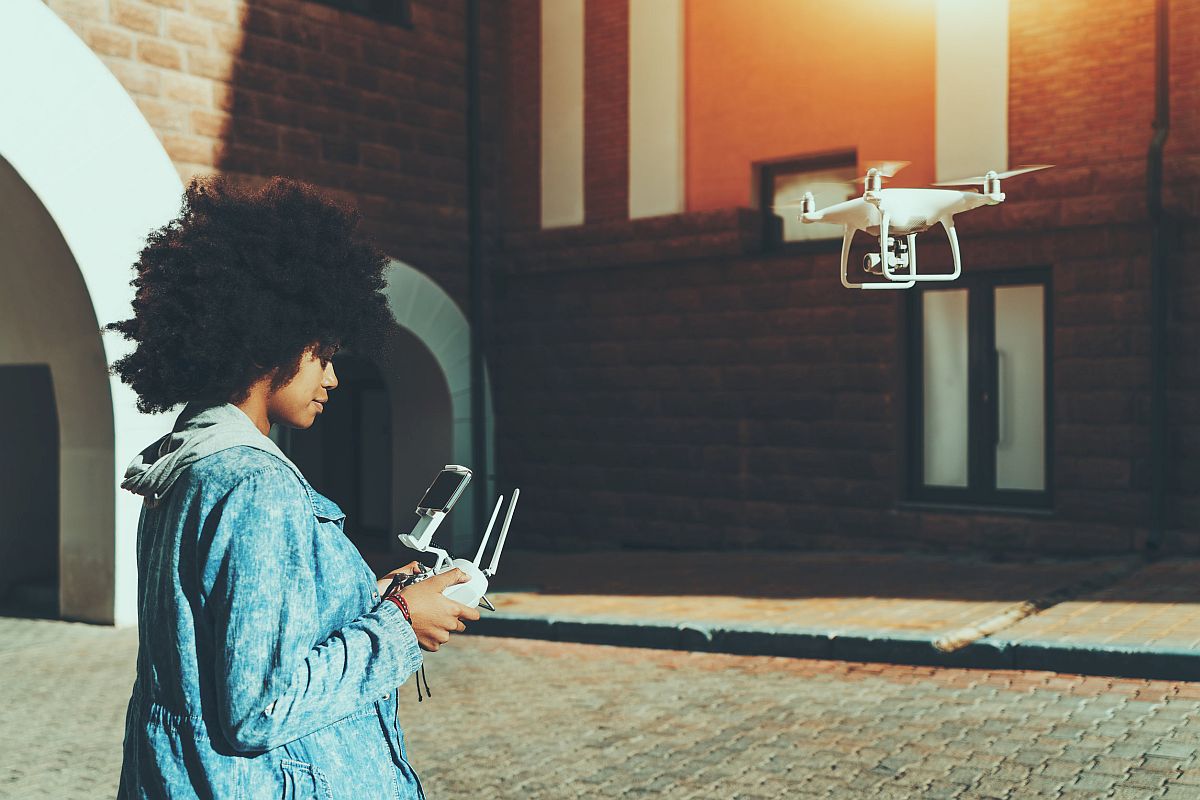 woman standing on pavement in urban settings and remotely tuning camera of flying drone; drones lesson plans concept