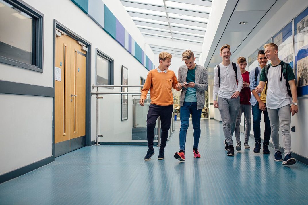 Group of teenage boys are walking down the school hall together to go for their lunch break. They are talking and laughing and some of the boys are using smart phones.