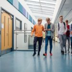 Group of teenage boys are walking down the school hall together to go for their lunch break. They are talking and laughing and some of the boys are using smart phones.