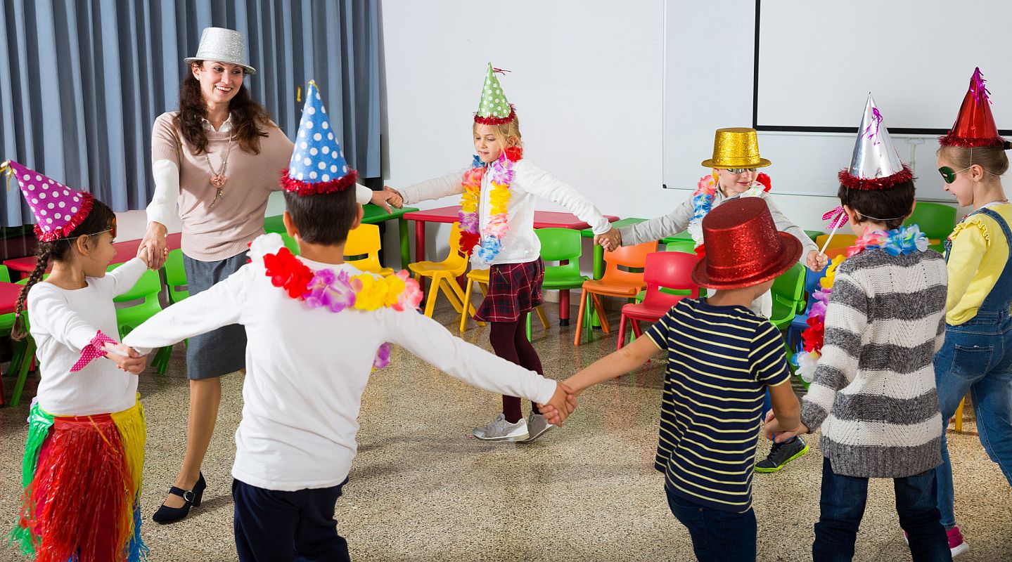 Happy kids and female teacher in funny hats and festive accessories dancing together in schoolroom; indoor recess concept