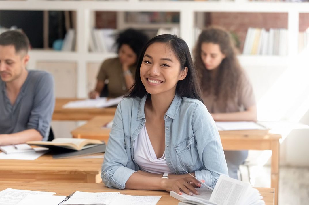 Smiling smart female asian teen girl looking at camera sitting at desk studying in classroom with diverse classmates group, happy chinese college university student in international class portrait
