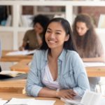 Smiling smart female asian teen girl looking at camera sitting at desk studying in classroom with diverse classmates group, happy chinese college university student in international class portrait