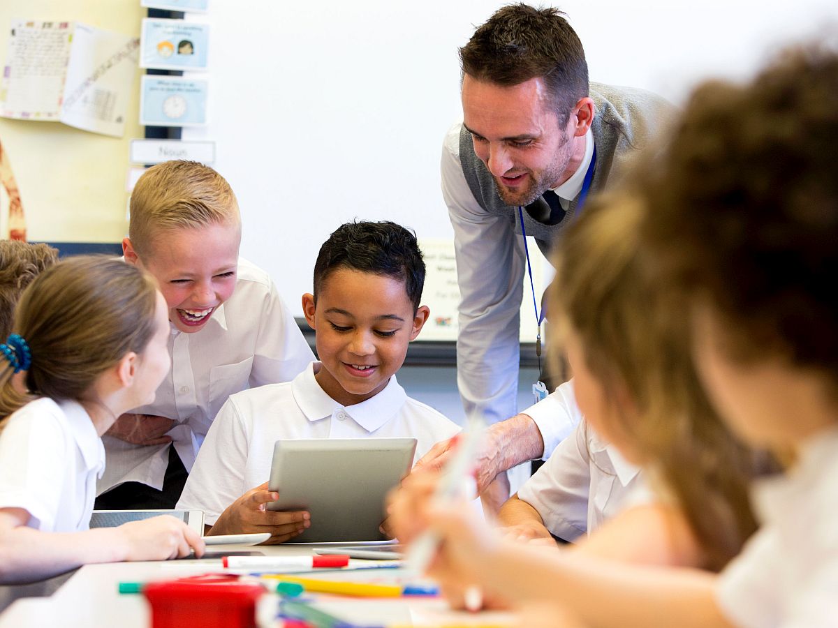 A male teacher sits supervising a group of children who are working on whiteboards and digital tablets; group projects concept