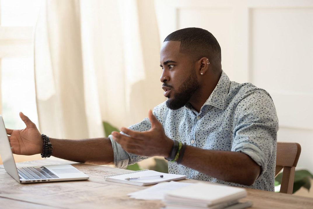 African guy sit at desk makes video call talks feels outraged discuss problem with person distantly, annoyed man has computer slow internet lost wifi connection unpleasant email too much spam concept