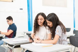 Smiling teenage friends looking at mobile phone while sitting in classroom