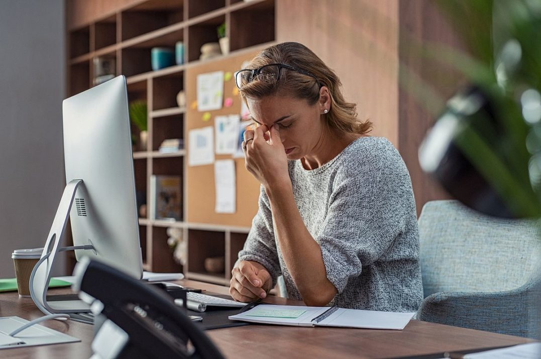 Exhausted businesswoman having a headache in modern office. Mature creative woman working at office desk with spectacles on head feeling tired. Stressed casual business woman feeling eye pain while overworking on desktop computer.