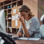 Exhausted businesswoman having a headache in modern office. Mature creative woman working at office desk with spectacles on head feeling tired. Stressed casual business woman feeling eye pain while overworking on desktop computer.