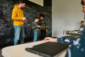 Young male science teacher standing near the blackboard, holding tablet pc and looking at his students while they are demonstrating their own robot vehicle. STEM disciplines, Math education concept