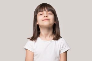 Close up head shot portrait image with relaxed little brown-haired girl, reduce stress. Wellbeing concept rest kid meditating and deep breathing on gray background, six year child with eyes closed