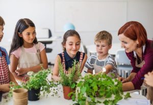 A group of young, happy students with teacher standing at the table in class, planting herbs; climate change lesson plans concept