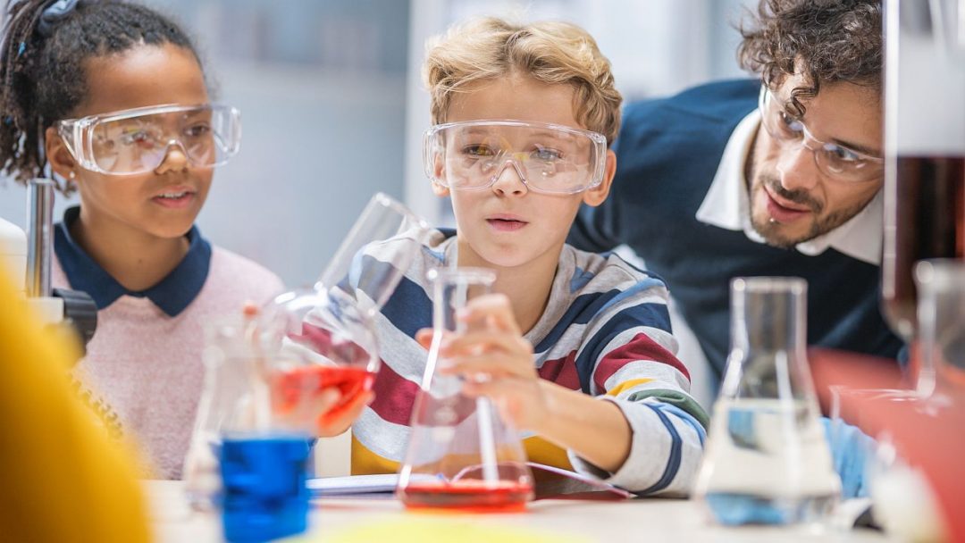 Young students mixing liquids in beakers, with teacher supervising; chemistry classroom concept