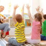 Teacher playing guitar with young students doing hand movements; add physical activity concept.