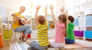 Teacher playing guitar with young students doing hand movements; add physical activity concept.