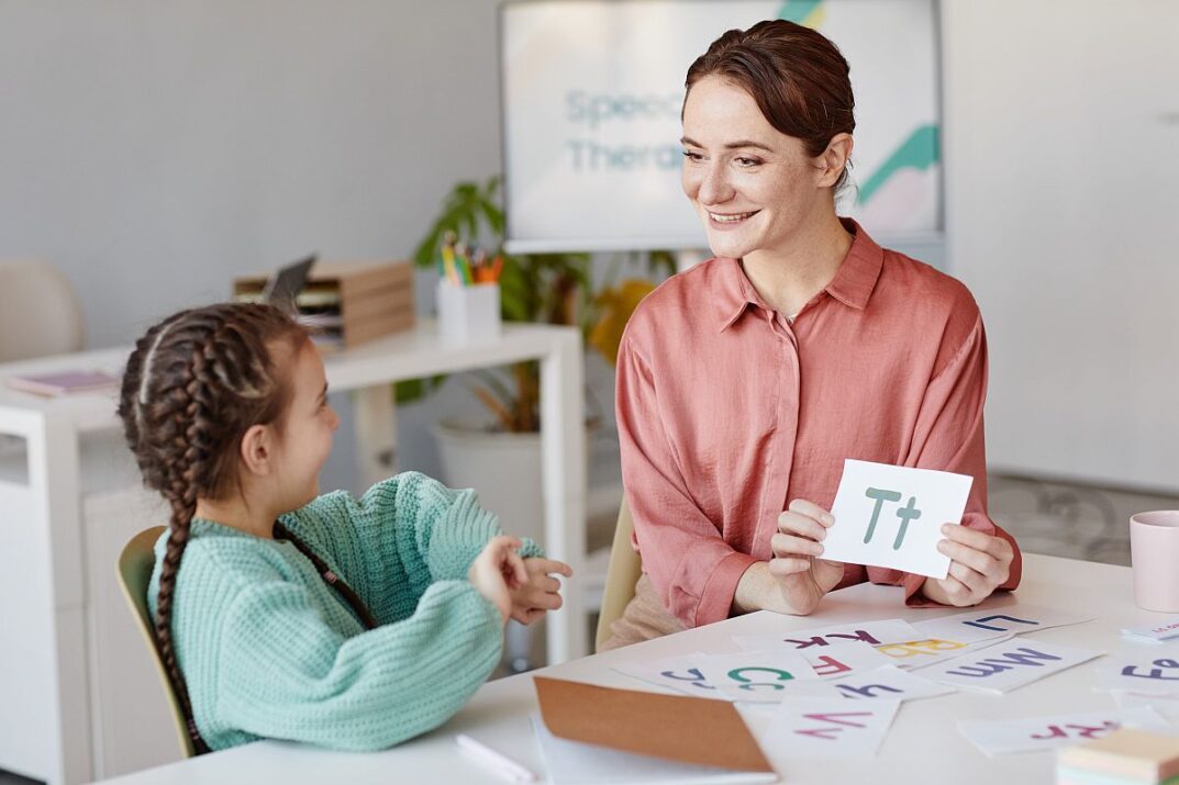 Woman showing alphabet card to young girl at table; reading specialists concept