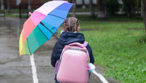 Young student with pink backpack and rainbow umbrella walking in the rain; weather lesson plans concept