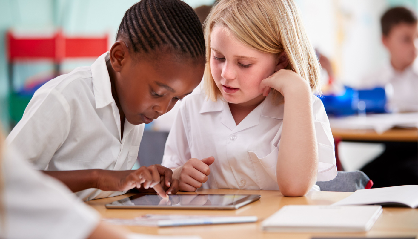 Two Elementary School Pupils Wearing Uniform Using Digital Tablet At Desk; AI lesson plans concept