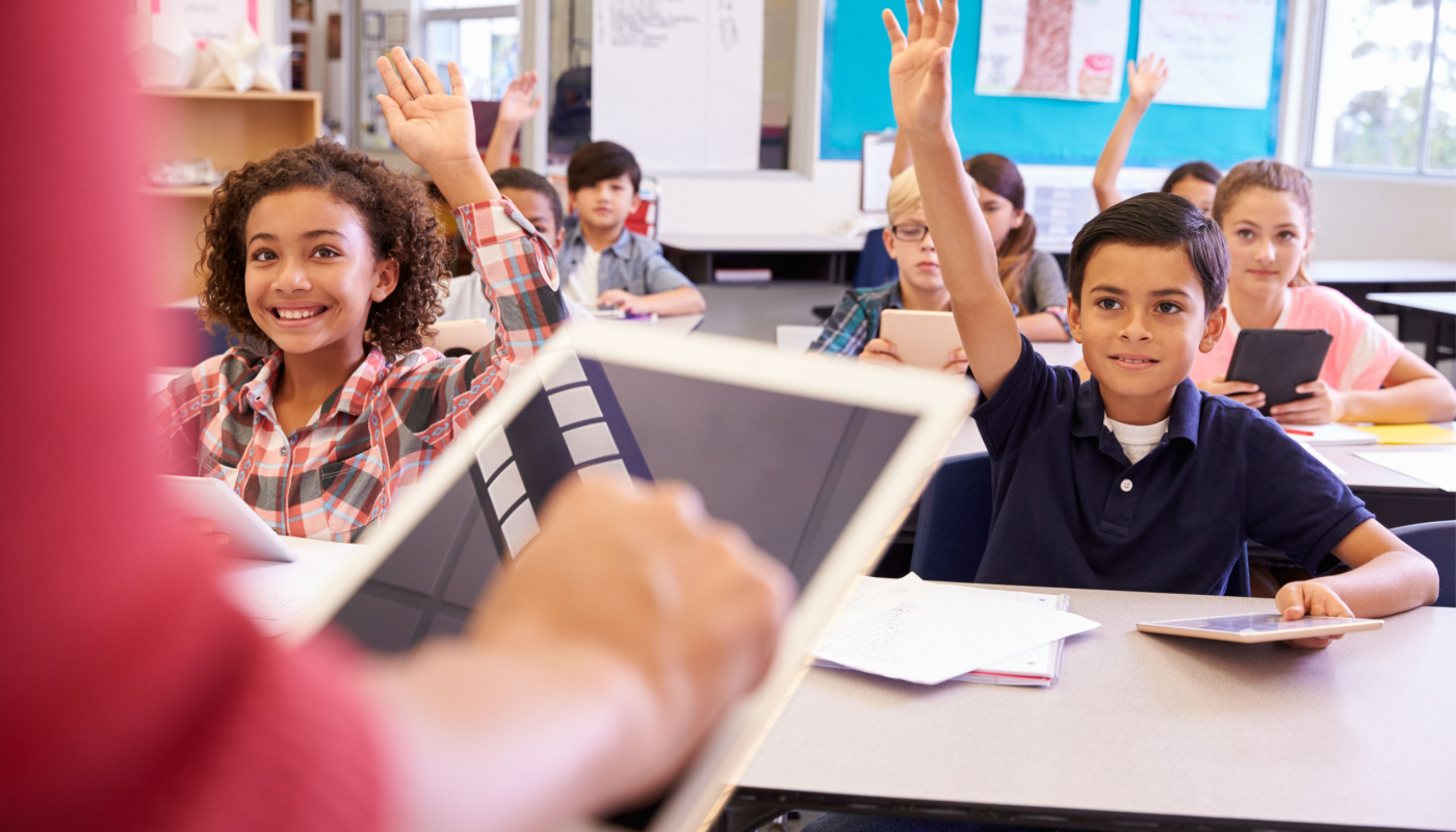 Teacher using tablet computer in elementary school lesson; kids raising hands; AI lesson plans concept