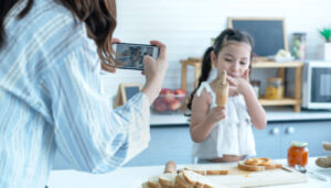 Mother taking pictures of her daughter or recording a vlog in her kitchen; internet privacy lessons concept