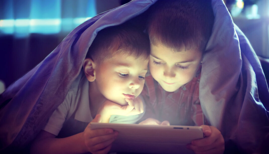 Two kids using tablet under a blanket at night; internet privacy lessons concept