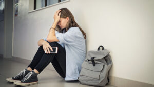 Upset teen holding smartphone and sitting on the school floor, head in hands; internet privacy lessons concept