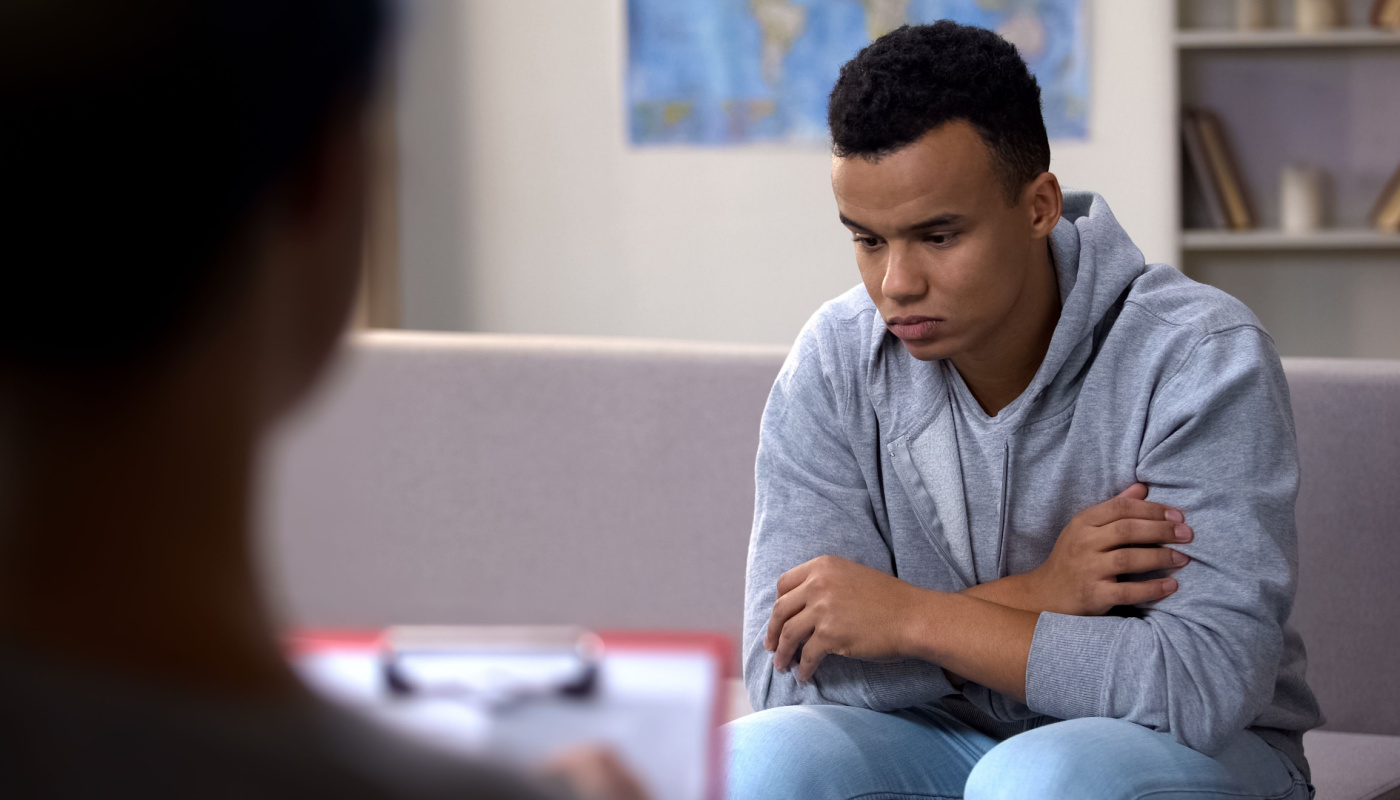 Depressed-looking teen seated on couch, opposite adult with clipboard; school psychologists concept